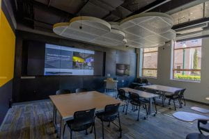 An image of the Social Media Lab. The wall of monitors is showing a website in a large format. There are three large table with chairs surrounding them.