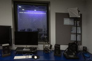 An image of the Edit Bay looking into the Podcast Studio. There is a desk with an iMac, mic, speakers and small TV.